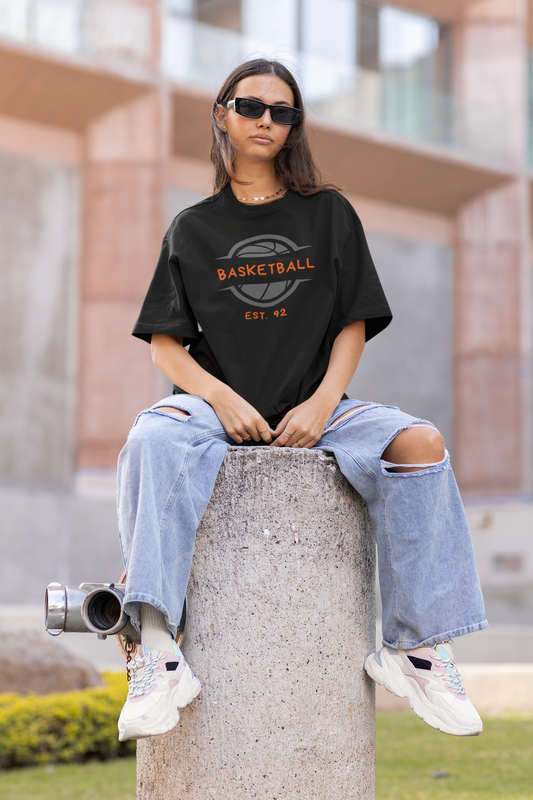 Mockup mit Model Schwarzes Oversized T-Shirt mit einem Print eines grauen Basketball-Emblems und dem orangefarbenen, stilisierten Text „BASKETBALL“ und „EST. 92“. 100% Baumwolle.