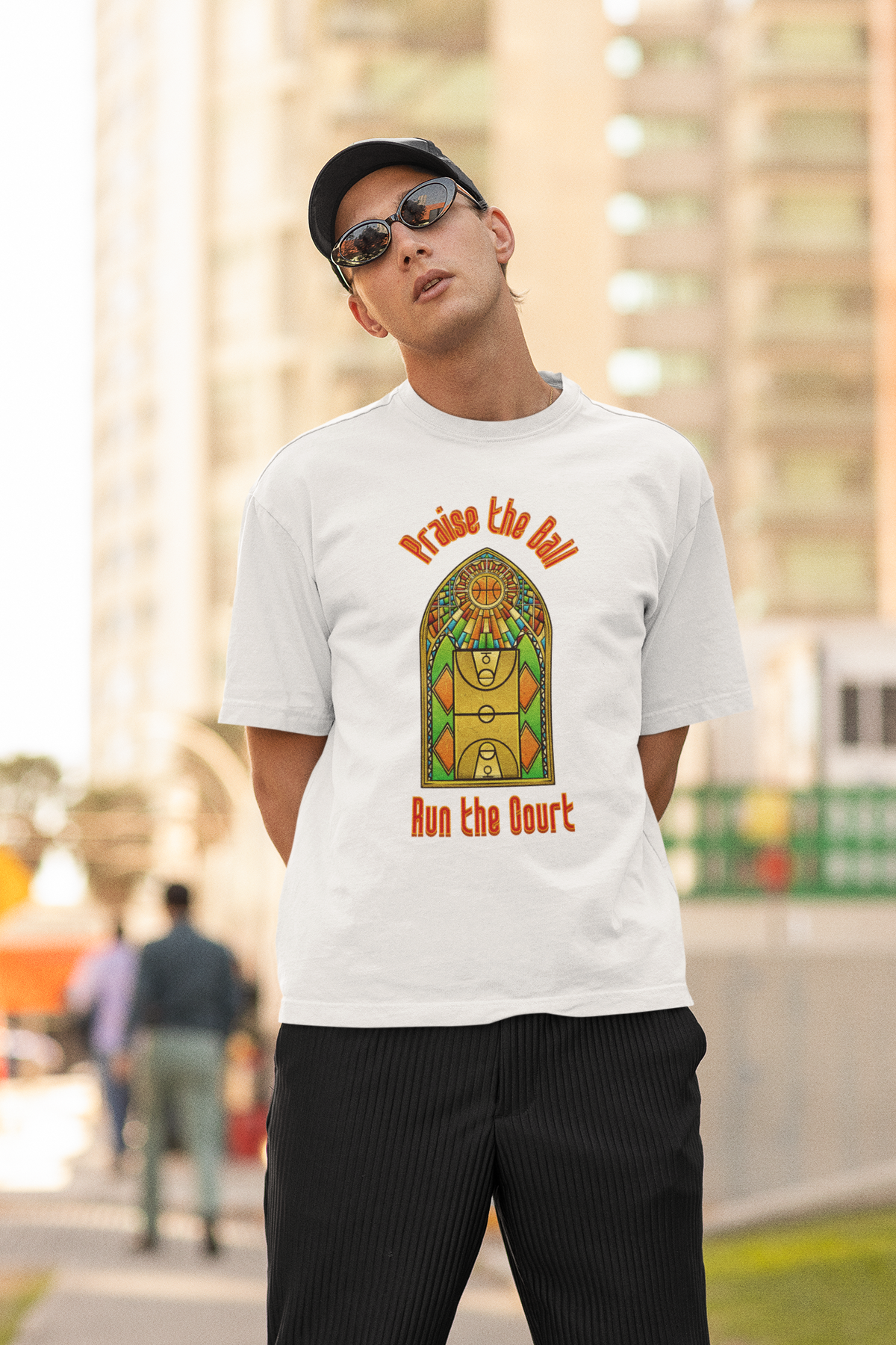 Mockup mit Model Weißes Oversized T-Shirt mit einem Print im Stil eines Kirchen-Buntglasfensters, der einen Basketball-Court zeigt. Darüber steht in Rot „Praise the Ball“ und darunter „Run the Court“. 100% Baumwolle.